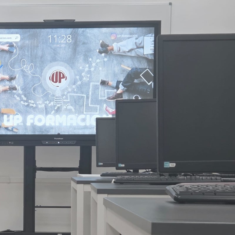 Aula tecnológica de UP Formación con ordenadores y pantalla interactiva mostrando contenidos formativos para cursos de informática y competencias digitales.